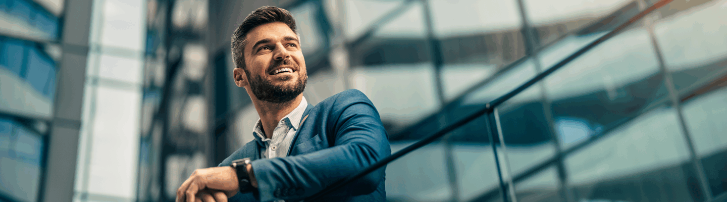 Mann in Anzug lehnt sich an ein Geländer. Er steht vor modernen Gebäuden.