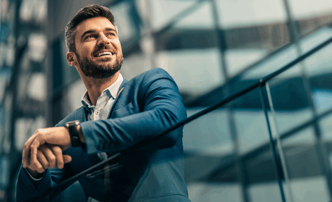 Mann in Anzug lehnt sich an ein Geländer. Er steht vor modernen Gebäuden.