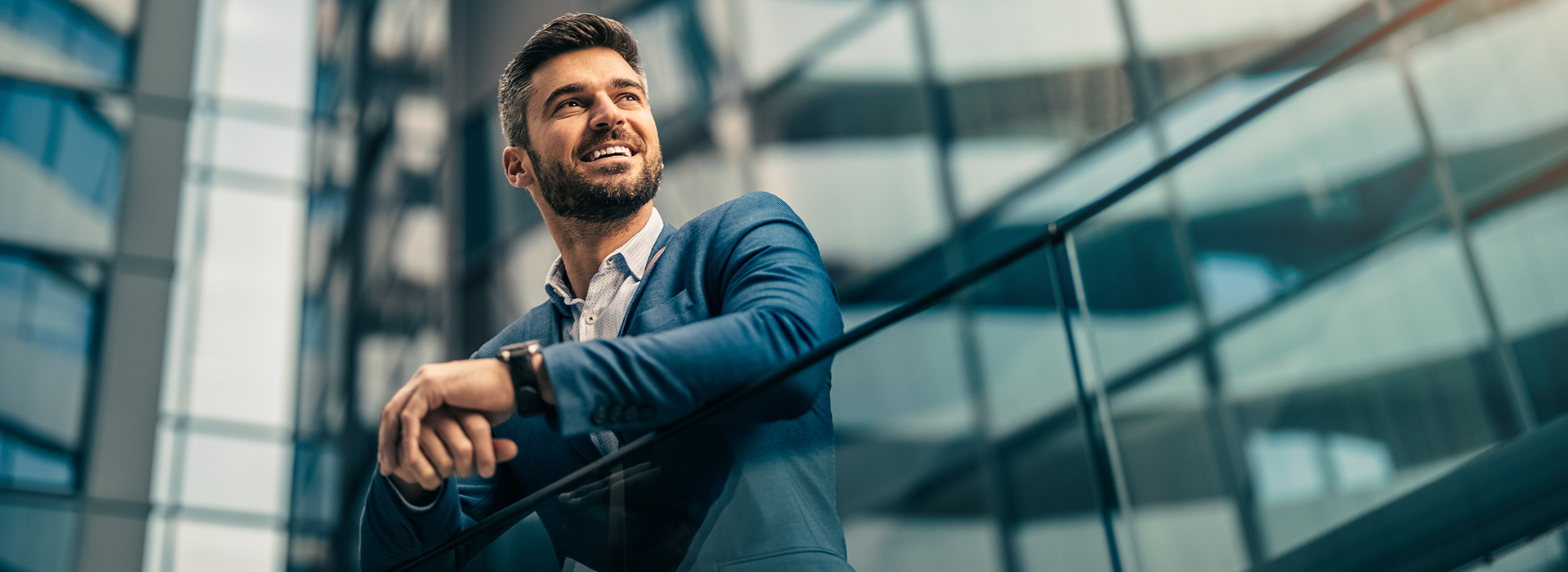Mann in Anzug lehnt sich an ein Geländer. Er steht vor modernen Gebäuden.