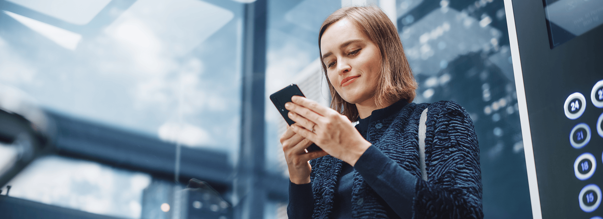 Frau steht in einem gläsernen Aufzug und schaut währenddessen auf ein Smartphone, das sie in den Händen hält