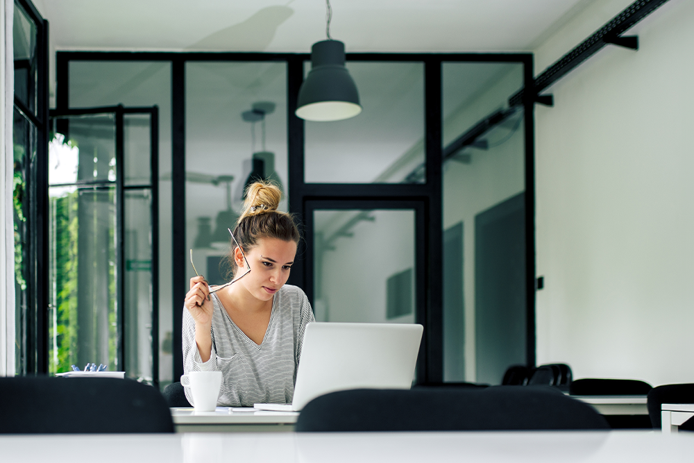 Eine Frau sitzt an einem Tisch mit geöffnetem Laptop vor ihr. In der Hand hält sie eine Brille während sie auf den Bildschirm schaut.