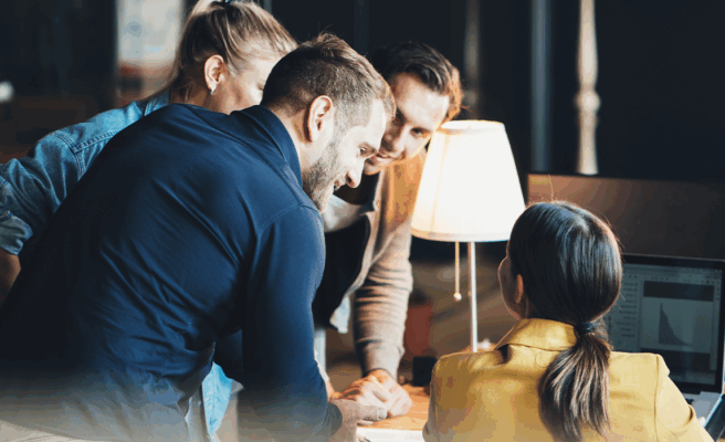 Personen, die um einen Schreibtisch herum stehen und sich unterhalten.