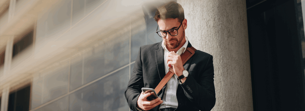 Mann, Business Casual, lehnt an einer Hauswand und schaut auf sein Handy - Brunata Minol Connect hilft bei der Fernablesung