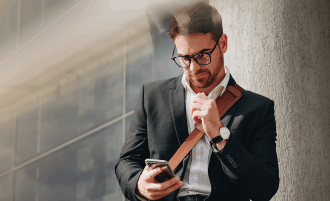 Mann, Business Casual, lehnt an einer Hauswand und schaut auf sein Handy - Brunata Minol Connect hilft bei der Fernablesung