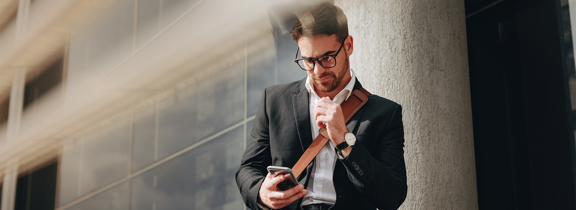 Mann, Business Casual, lehnt an einer Hauswand und schaut auf sein Handy - Brunata Minol Connect hilft bei der Fernablesung