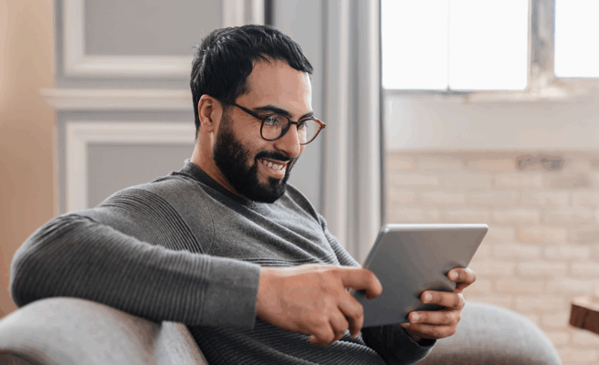 Mann sitzt auf einem Sofa und schaut etwas auf seinem Tablet an, das er in den Händen hält.
