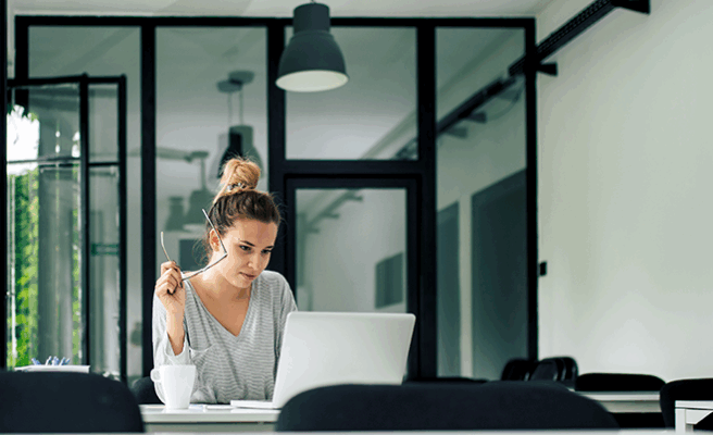 Eine Frau sitzt an einem Tisch mit geöffnetem Laptop vor ihr. In der Hand hält sie eine Brille während sie auf den Bildschirm schaut.