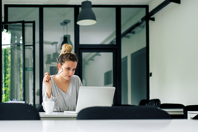 Eine Frau sitzt an einem Tisch mit geöffnetem Laptop vor ihr. In der Hand hält sie eine Brille während sie auf den Bildschirm schaut.