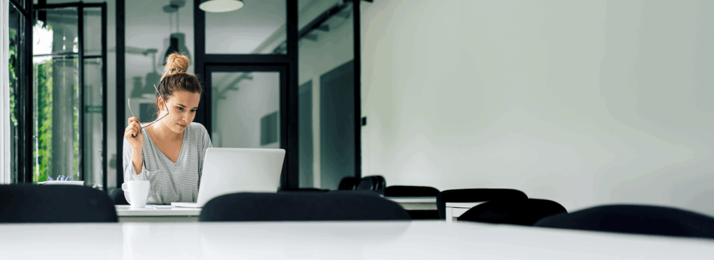 Eine Frau sitzt an einem Tisch mit geöffnetem Laptop vor ihr. In der Hand hält sie eine Brille während sie auf den Bildschirm schaut.