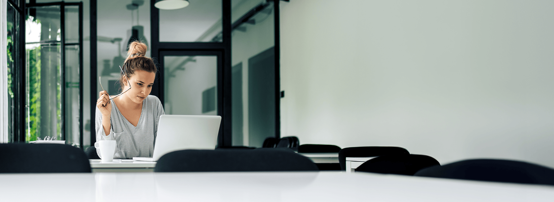 Eine Frau sitzt an einem Tisch mit geöffnetem Laptop vor ihr. In der Hand hält sie eine Brille während sie auf den Bildschirm schaut.