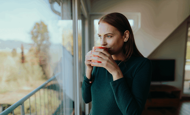 Frau hält einen Kaffeebecher in den Händen vor ihrem Gesicht und schaut aus einem Fenster