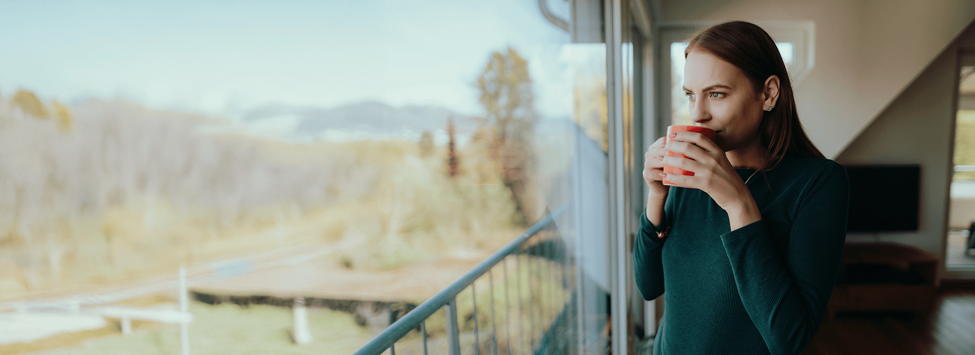 Frau hält einen Kaffeebecher in den Händen vor ihrem Gesicht und schaut aus einem Fenster