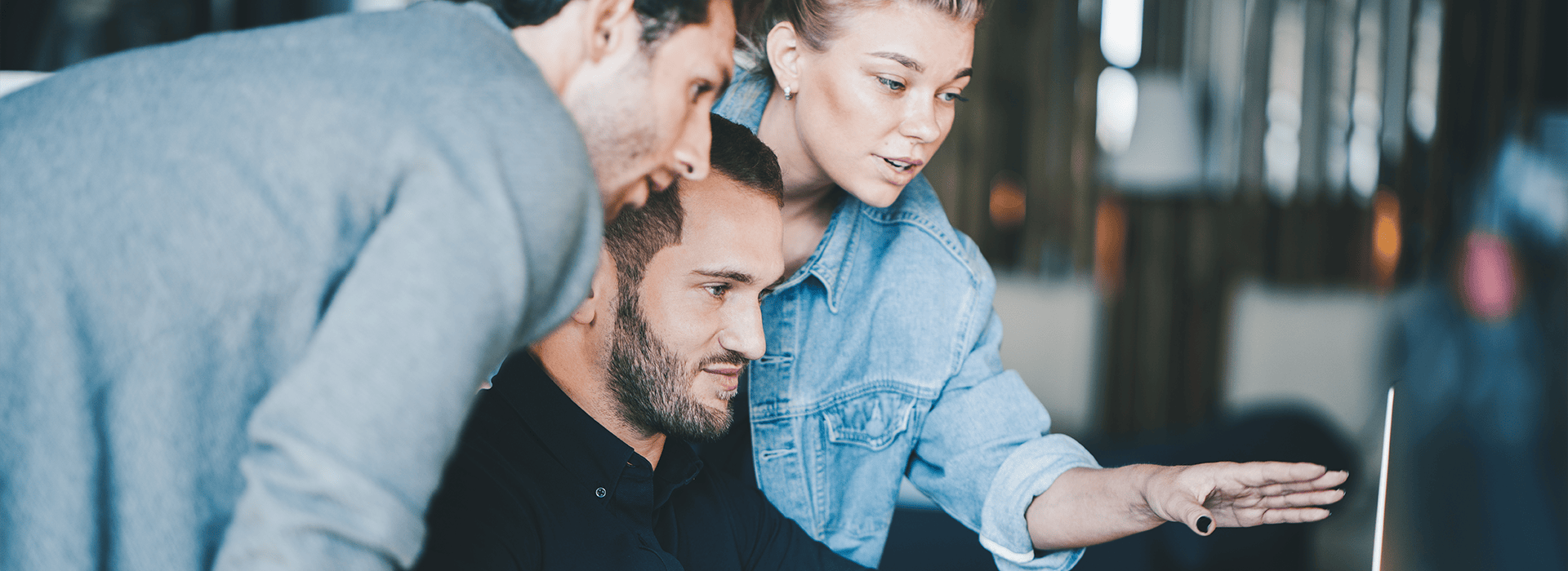 Drei Personen schauen auf einen Bildschirm an einem Schreibtisch. Die Frau zeigt auf etwas mit ihrer Hand.