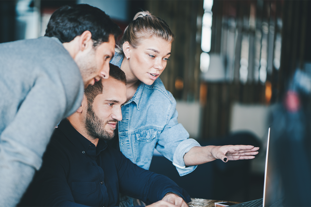 Drei Personen schauen auf einen Bildschirm an einem Schreibtisch. Die Frau zeigt auf etwas mit ihrer Hand.