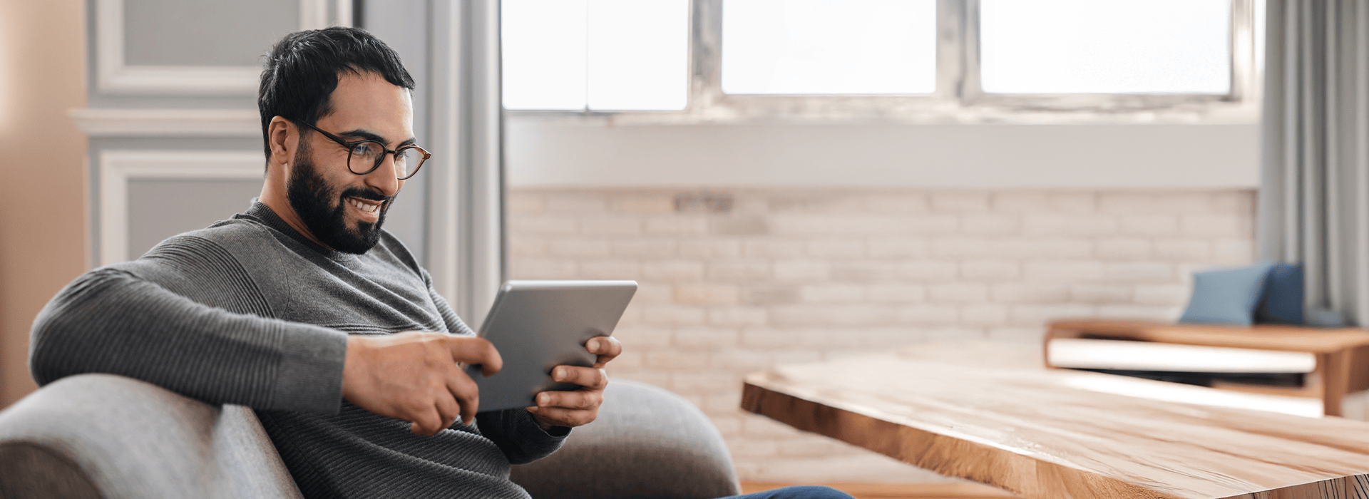 Mann sitzt auf einem Sofa und schaut etwas auf seinem Tablet an, das er in den Händen hält.