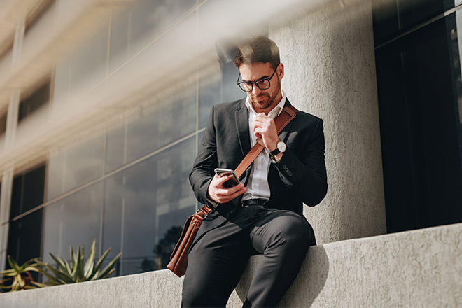 Mann, Business Casual, lehnt an einer Hauswand und schaut auf sein Handy - Brunata Minol Connect hilft bei der Fernablesung