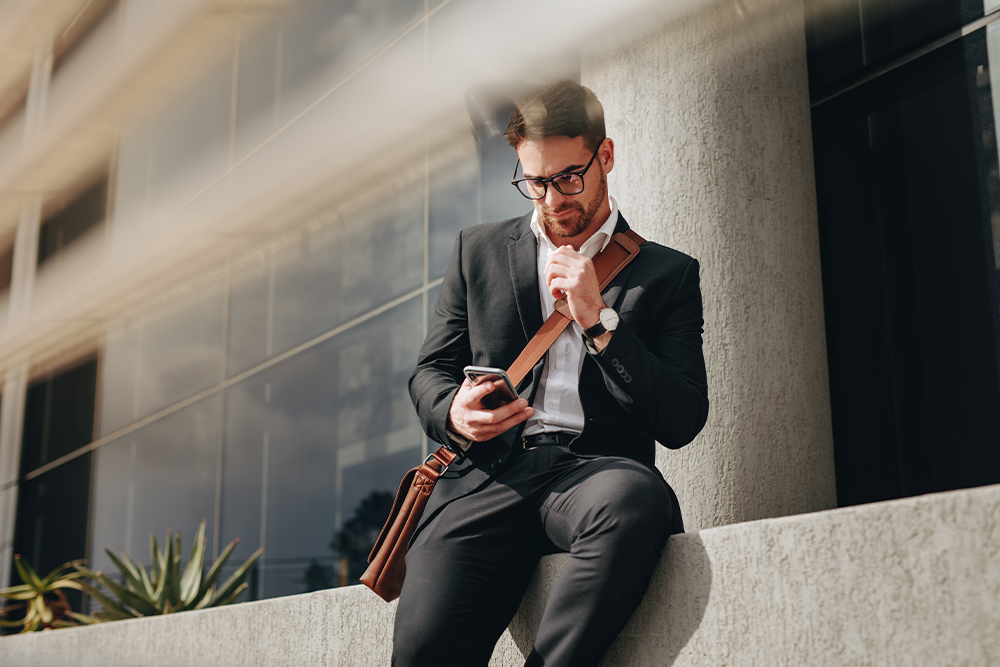 Mann, Business Casual, lehnt an einer Hauswand und schaut auf sein Handy - Brunata Minol Connect hilft bei der Fernablesung