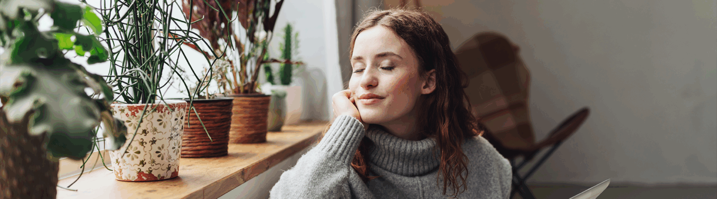 Frau mit geschlossenen Augen und entspanntem Gesichtsausdruck lehnt an einer Heizung, sie hält ein Tablet in der Hand.