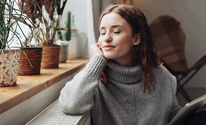 Frau mit geschlossenen Augen und entspanntem Gesichtsausdruck lehnt an einer Heizung, sie hält ein Tablet in der Hand.