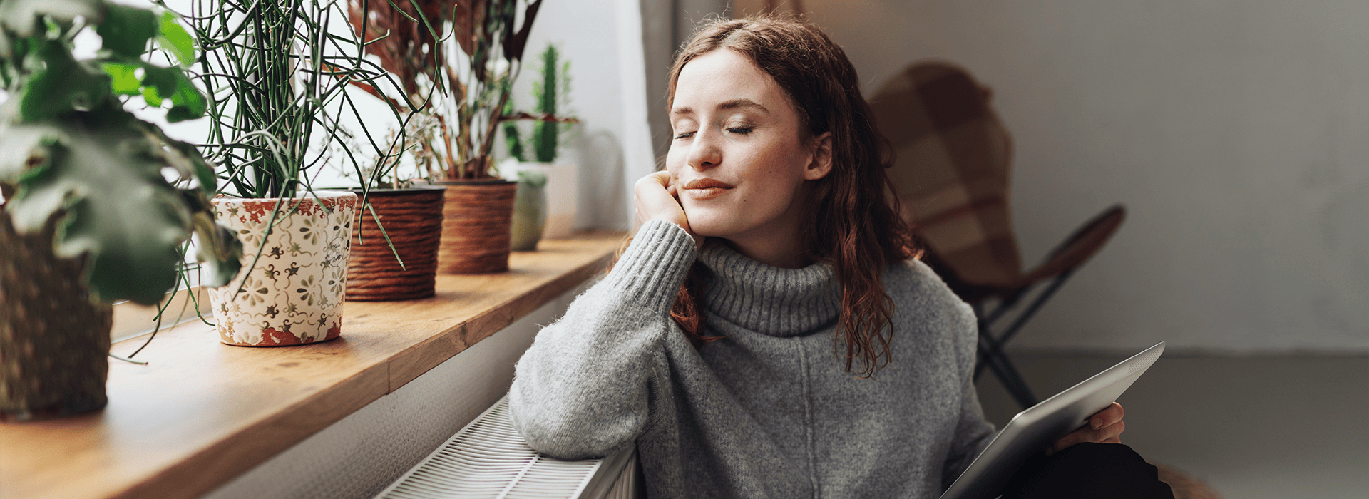 Frau mit geschlossenen Augen und entspanntem Gesichtsausdruck lehnt an einer Heizung, sie hält ein Tablet in der Hand.